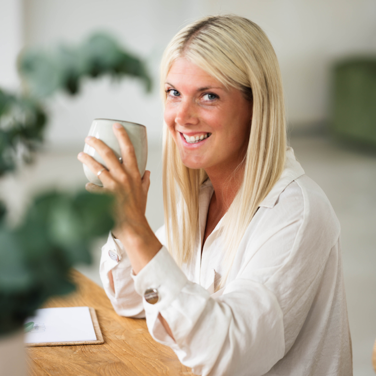 Blonde Frau mit einer Tasse in der Hand am Schreibtisch. Sie lächelt.